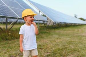 ein glücklich wenig Junge im ein Gelb Helm ist Stehen auf ein Solar- Panel Bauernhof. foto
