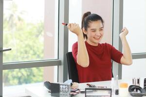 schön asiatisch Frau Blogger lächelnd und Hand halten rot Lippenstift zum machen oben Lernprogramm im Kosmetika Webseite. Marketing im Sozial Medien Konzept. foto