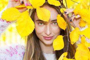 Schönes Mädchen, das ihr Gesicht hinter einem Herbstblatt versteckt, fröhliche Frau versteckt das Gesicht des gelben Herbstblattes, das Nahaufnahme trägt, Herbstkonzept foto