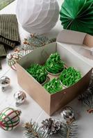Weihnachtsbaumförmige Cupcakes in handwerklicher Öko-Box, umgeben von festlicher Dekoration foto