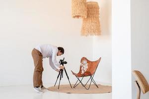 männlicher Fotograf, der in minimalem, hellem und luftigem Interieur arbeitet, weißer und beigefarbener Stuhl, Teppich und Kissen foto