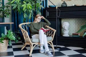 junge blonde Frau sitzt in einem bequemen Stuhl und liest ein Buch