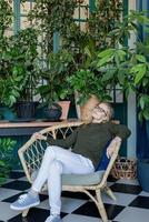 junge blonde Frau sitzt in einem bequemen Stuhl, umgeben von Pflanzen