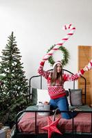 junge Frau in rotem Pullover und Weihnachtsmütze tanzt auf dem Bett und feiert Weihnachten foto
