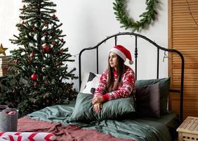 junge Frau in rotem Pullover und Weihnachtsmütze sitzt auf dem Bett und feiert Weihnachten foto