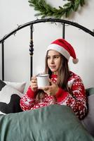junge Frau in rotem Pullover und Weihnachtsmütze sitzt auf dem Bett und feiert Weihnachten mit einer Tasse heißem Getränk foto