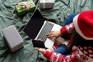Frau in Weihnachtskleidung, die mit Laptop und Handy auf dem Bett sitzt foto