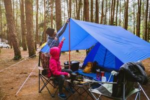 Junge asiatische Paare, die das Camping im Kiefernwald genießen, sitzen und essen auf der Campingseite inmitten der Natur. foto