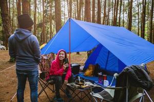 Junge asiatische Paare, die das Camping im Kiefernwald genießen, sitzen und essen auf der Campingseite inmitten der Natur.