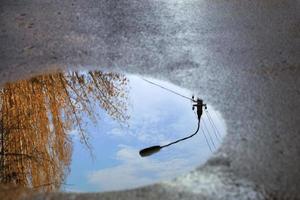 Reflexion des Himmels, die Silhouette einer Straßenlaterne und ein Baum sonnenbeschienen in einer Pfütze auf Asphalt. foto
