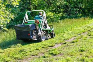 Ein erfahrener Gärtner mäht hohes Gras auf einem Traktor-Rasenmäher, während er den Hang eines Parkhains erklimmt, Kopienraum. foto