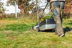 Ein Gärtner mäht grünes Gras mit einem elektrischen Rasenmäher in seinem Hinterhof an einem sonnigen Herbsttag, Rückansicht. foto