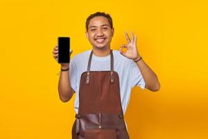 Fröhlicher asiatischer Mann, der einen leeren Bildschirm des Smartphones zeigt und ein Okay-Zeichen auf gelbem Hintergrund gestikuliert