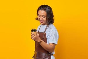 Porträt eines gutaussehenden Mannes mit Schürze, der auf eine Tasse Kaffee schaut, die isoliert auf gelbem Hintergrund gehalten wird foto