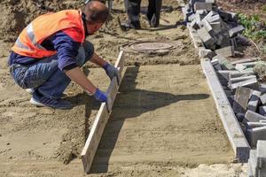 der arbeiter räumt und richtet den sandsockel mit einem holzbrett aus, um anschließend die fliesen auf dem gehweg und um den kanalschacht zu verlegen. foto