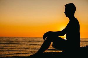 Silhouette eines Mannes, der am Strand auf Meereshintergrund und Sonnenuntergang sitzt und nachdenkt foto