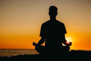 Silhouette eines Mannes in Meditationshaltung am Strand auf Meereshintergrund und Sonnenuntergang foto