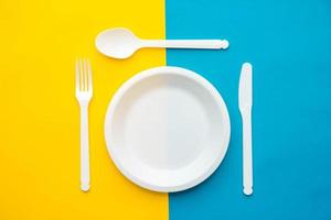 weiße Gabel, Messer, Löffel und Teller aus Kunststoff auf gelbem und blauem Hintergrund