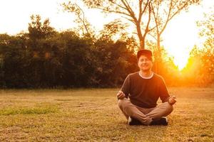 Mann in meditativer Position sitzt auf einer Wiese vor dem Hintergrund von Wald und Sonnenuntergang foto