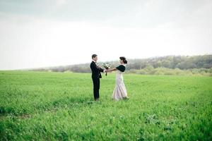 der Bräutigam im braunen Anzug und die Braut im elfenbeinfarbenen Kleid auf grüner Wiese foto