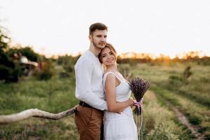 Mädchen in einem weißen Sommerkleid und ein Typ in einem weißen Hemd auf einem Spaziergang bei Sonnenuntergang mit einem Blumenstrauß foto