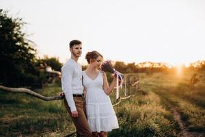 Mädchen in einem weißen Sommerkleid und ein Typ in einem weißen Hemd auf einem Spaziergang bei Sonnenuntergang mit einem Blumenstrauß foto