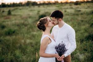 Mädchen in einem weißen Sommerkleid und ein Typ in einem weißen Hemd auf einem Spaziergang bei Sonnenuntergang mit einem Blumenstrauß foto