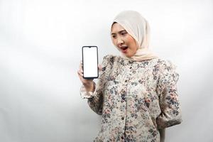 schöne junge asiatische muslimische frau schockiert, überrascht, wow-ausdruck, hand hält smartphone mit weißem oder leerem bildschirm, fördert app, fördert produkt, präsentiert etwas, isoliert foto