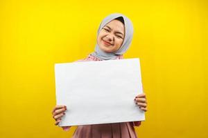 hübsche junge muslimische Frau, die fröhliche leere leere Banner, Plakate, weiße Tafeln, leere Schilder, weiße Werbetafeln hält, etwas im Kopierraum präsentiert, Werbung foto
