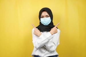 muslimische Frau mit medizinischer Maske, Hand zeigt auf leeren Raum, Hand zeigt nach oben und präsentiert etwas, isoliert auf gelbem Hintergrund