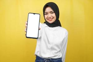 schöne junge asiatische muslimische frau, die selbstbewusst, enthusiastisch und fröhlich lächelt, mit den händen, die das smartphone halten, die anwendung fördern, etwas fördern, einzeln auf gelbem hintergrund foto