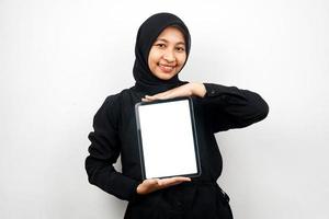schöne junge asiatische muslimische frau, die lächelt, hand hält tablette mit weißem oder leerem bildschirm, fördert app, fördert produkt, präsentiert etwas, aufgeregt und fröhlich, einzeln auf weißem hintergrund foto