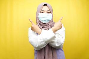 muslimische Frau mit medizinischer Maske, Hand zeigt auf leeren Raum, Hand zeigt nach oben und präsentiert etwas, isoliert auf gelbem Hintergrund