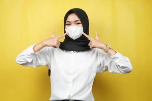 muslimische frau mit weißer maske, mit der hand, die auf maske zeigt, koronavirus-geste verhindern, covid-19 verhindern, isoliert auf gelbem hintergrund foto