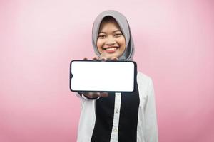 schöne junge asiatische muslimische frau, die selbstbewusst, enthusiastisch und fröhlich lächelt, mit den händen, die smartphone halten, anwendung präsentieren, spiel präsentieren, einzeln auf rosa hintergrund, werbekonzept foto