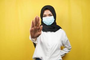 muslimische Frau mit medizinischer Maske, mit Stoppschild-Hand, Ablehnungsschild-Hand, Abstandsschild-Hand nicht halten, Handzeichen nicht nähern, isoliert auf gelbem Hintergrund