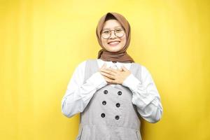 schöne junge asiatische muslimische frau, die überrascht und fröhlich lächelt, mit den händen, die die brust halten, aufgeregt, nicht erwartet, die kamera einzeln auf gelbem hintergrund betrachtend