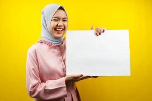 hübsche junge muslimische Frau, die fröhliche leere leere Banner, Plakate, weiße Tafeln, leere Schilder, weiße Werbetafeln hält, etwas im Kopierraum präsentiert, Werbung foto