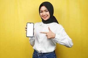 schöne junge asiatische muslimische frau, die selbstbewusst, enthusiastisch und fröhlich lächelt, mit der hand, die auf das smartphone zeigt, etwas fördert, die hand fördert die app, einzeln auf gelbem hintergrund foto