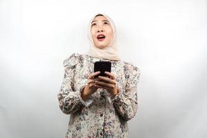 schöne junge asiatische muslimische frau schockiert, überrascht, wow-ausdruck, hand, die smartphone hält, nach oben schauend, isoliert auf weißem hintergrund
