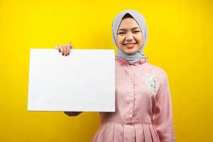 hübsche junge muslimische Frau, die fröhliche leere leere Banner, Plakate, weiße Tafeln, leere Schilder, weiße Werbetafeln hält, etwas im Kopierraum präsentiert, Werbung foto