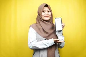 Schöne junge asiatische muslimische Frau, die selbstbewusst und aufgeregt mit den Händen lächelt, die Smartphone halten, App bewerben, etwas fördern, isoliert auf gelbem Hintergrund, Werbekonzept
