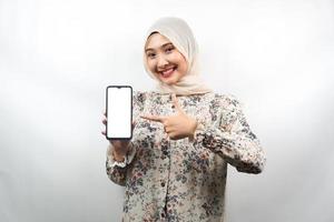 Schöne junge asiatische muslimische Frau, die selbstbewusst, enthusiastisch und fröhlich lächelt, mit der Hand auf das Smartphone zeigt, etwas fördert, die Hand App fördert, isoliert auf weißem Hintergrund foto