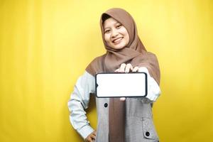 schöne junge asiatische muslimische frau, die selbstbewusst, enthusiastisch und fröhlich lächelt, mit den händen, die das smartphone halten, die anwendung fördern, das spiel fördern, einzeln auf gelbem hintergrund, werbekonzept