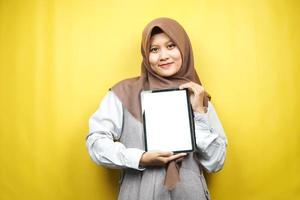 Schöne junge asiatische muslimische Frau lächelnd, aufgeregt und fröhlich, die Tablette mit weißem oder leerem Bildschirm hält, App fördert, Produkt fördert, etwas präsentiert, isoliert auf gelbem Hintergrund foto