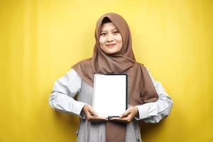 Schöne junge asiatische muslimische Frau lächelnd, aufgeregt und fröhlich, die Tablette mit weißem oder leerem Bildschirm hält, App fördert, Produkt fördert, etwas präsentiert, isoliert auf gelbem Hintergrund foto
