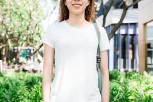 Asiatisches Hipster-Mädchen mit langen braunen Haaren in weißem leerem T-Shirt steht mitten auf der Straße. Eine Frau in Streetwear steht auf einem grünen urbanen Hintergrund. leerer Mock-up-Platz für Text oder Design. foto