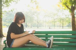 Happy Hipster junge asiatische Frau in ihr Tagebuch im Park schreiben. glückliche Hipster junge asiatische Frau, die an Notebook im Park arbeitet. Student, der im Freien studiert. Lifestyle Frau Outdoor-Konzept. flackerndes Licht. foto