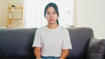 Depressive weinende asiatische Geschäftsfrau, die mit Kopfschmerzen gestresst ist und auf dem Sofa im Wohnzimmer des Hauses sitzt, wenn soziale Distanzierung zu Hause bleibt und Selbstquarantänezeit, Coronavirus-Konzept.