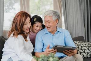 asiatische Großeltern und Enkelin mit Tablet zu Hause. Senior Chinese, Opa und Oma verbringen glücklich Zeit mit der Familie, entspannen sich mit jungen Mädchen, die soziale Medien überprüfen und auf dem Sofa im Wohnzimmerkonzept liegen foto